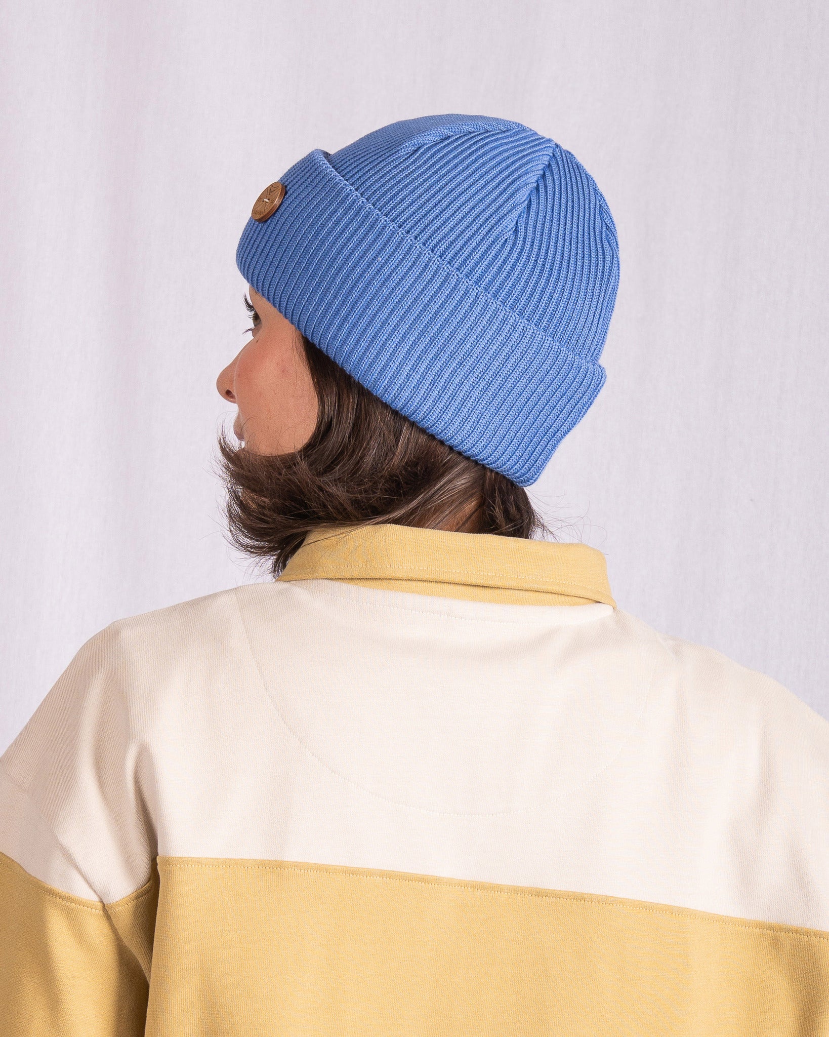 Person wearing a blue knit beanie and a beige jacket, facing away from the camera against a white background.