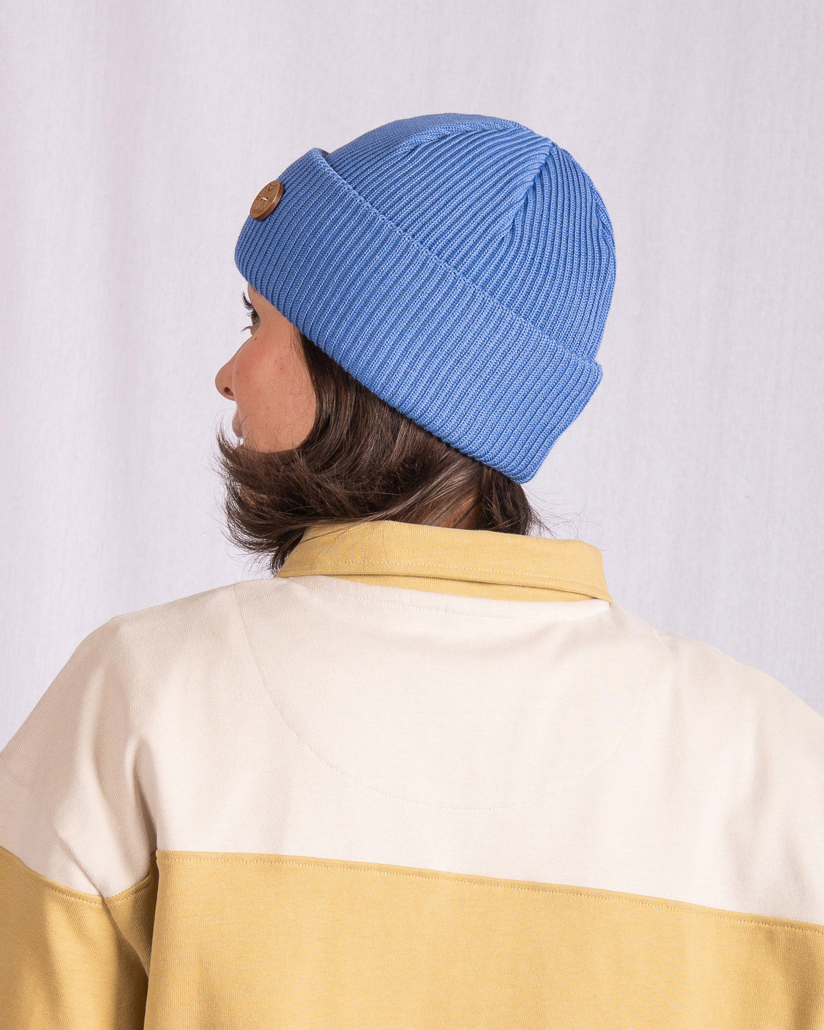 Person wearing a blue knit beanie and a beige jacket, facing away from the camera against a white background.