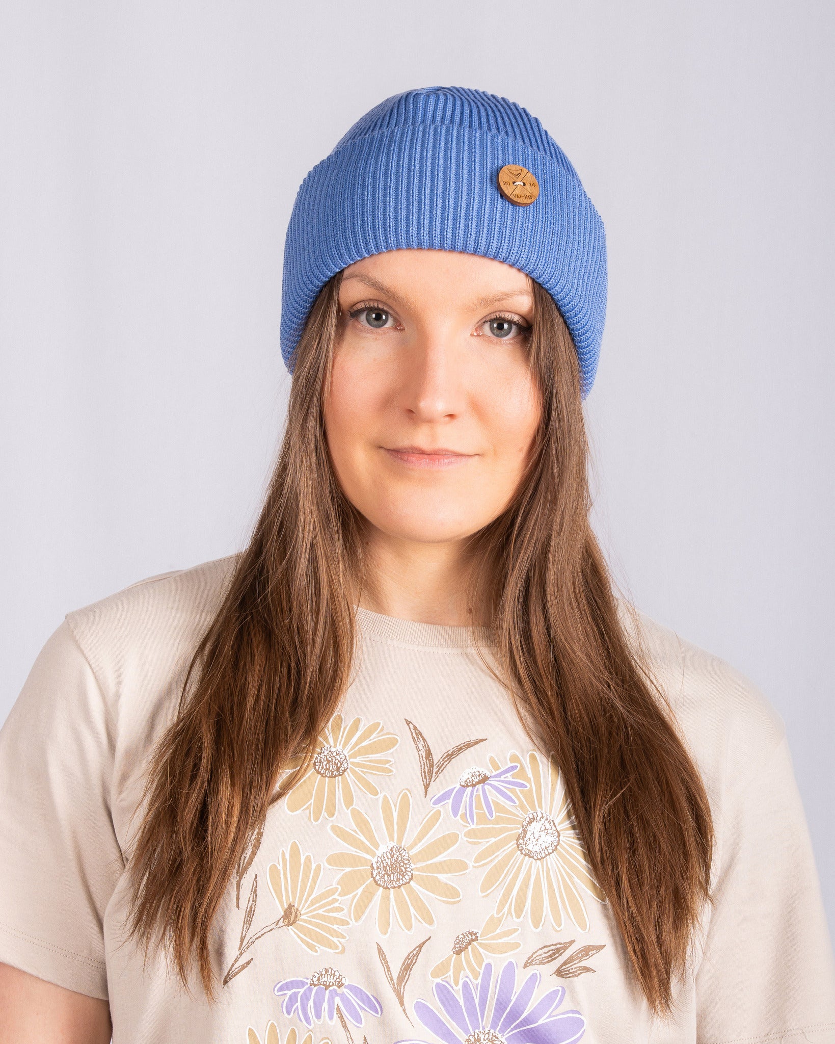 Woman wearing a blue knit beanie and beige shirt with a flower print, standing against a plain background.