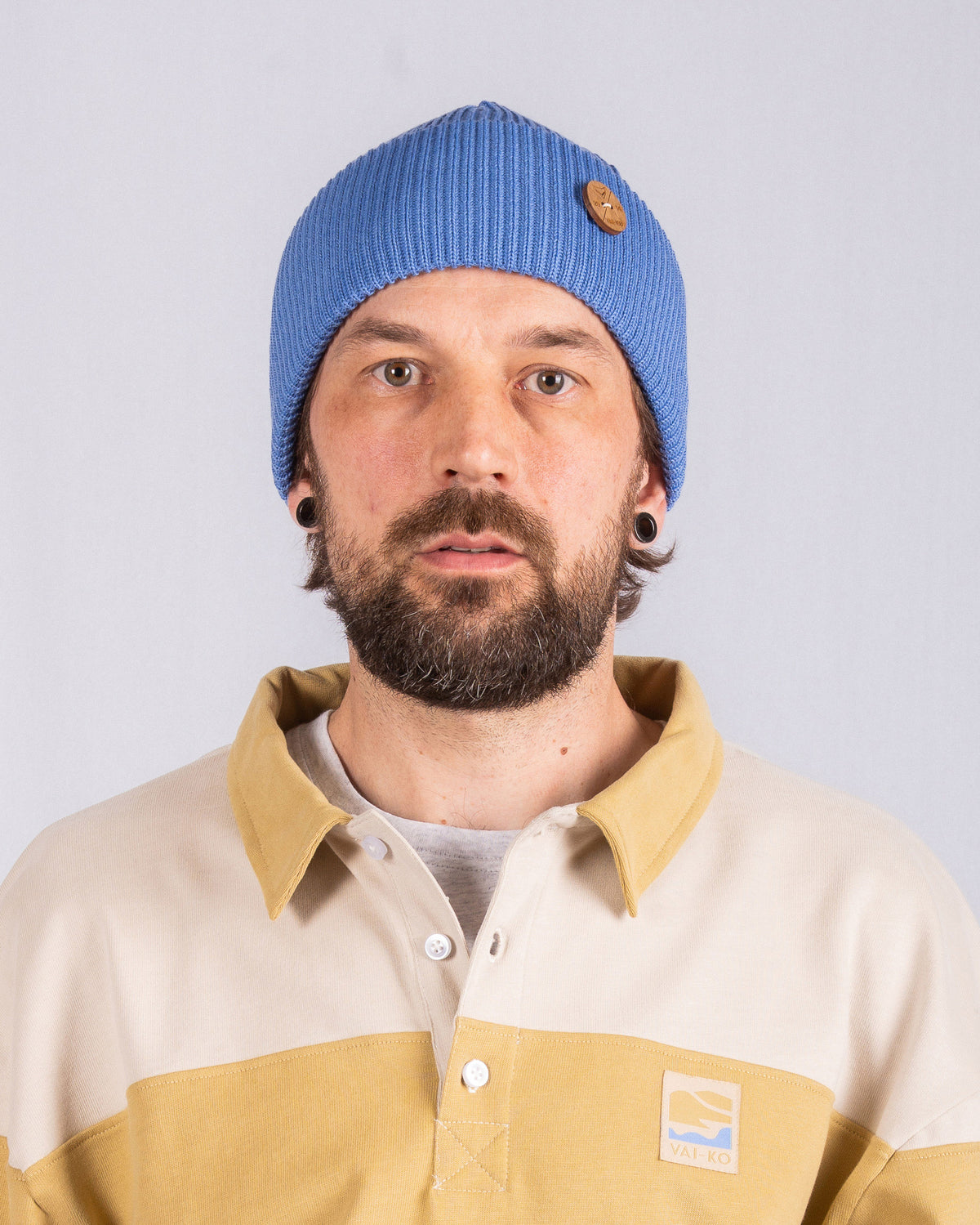 Man with a beard wearing a blue beanie and a beige collared shirt, facing the camera against a plain background.
