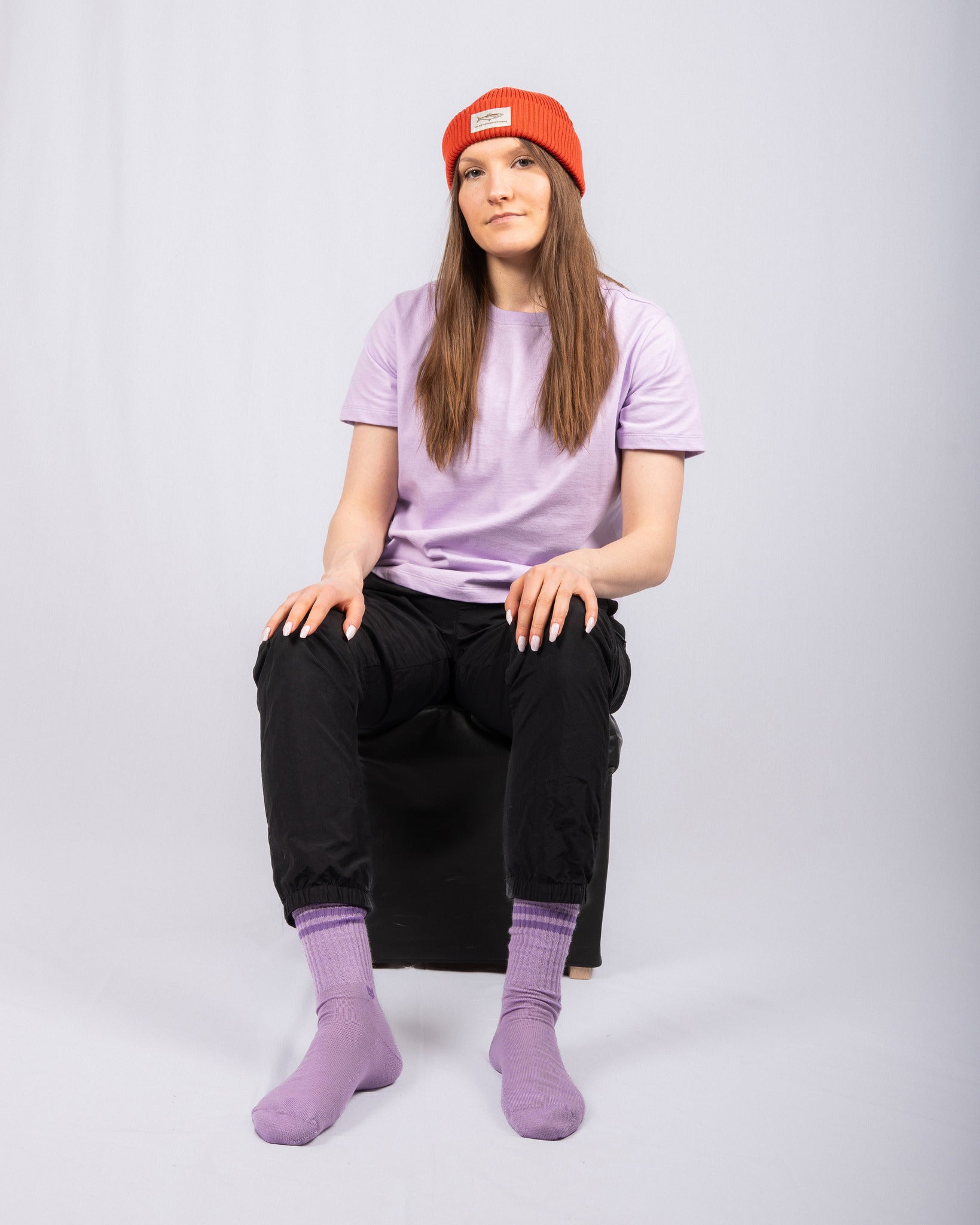 Person in lilac shirt, black pants, purple socks, and red beanie sits on a stool against a plain backdrop.