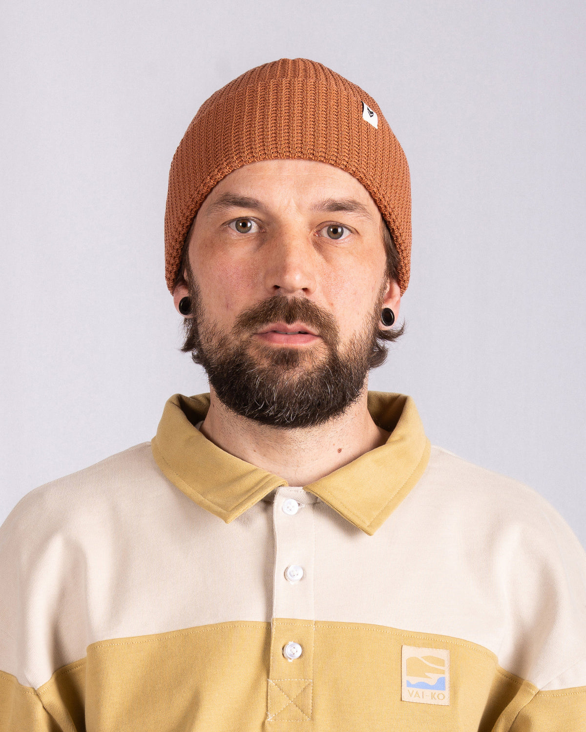 Man with a brown knit beanie, beige shirt with collar, and beard, facing forward against plain background.