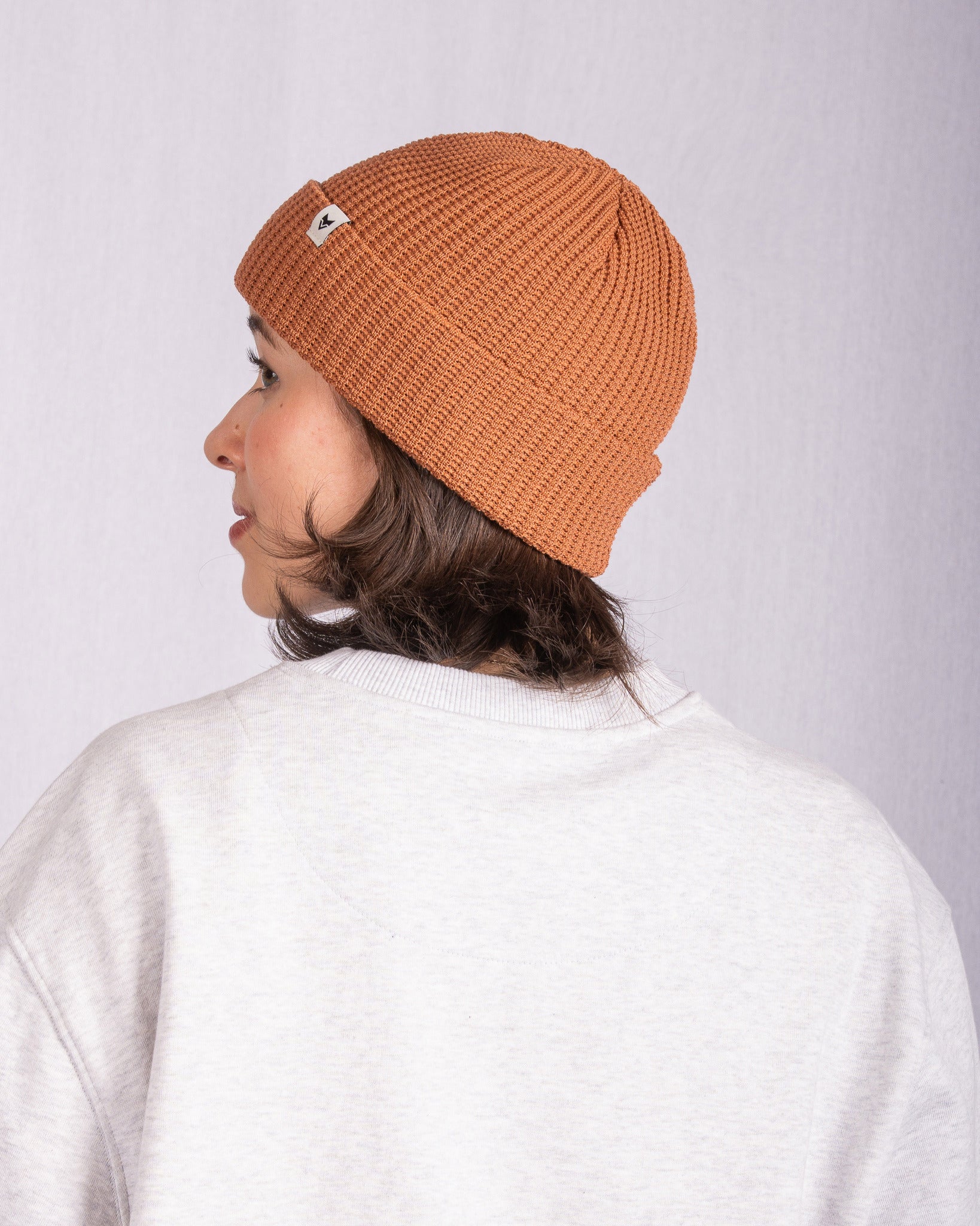 Person in a tan knit beanie and light sweater facing away, with a neutral light background.