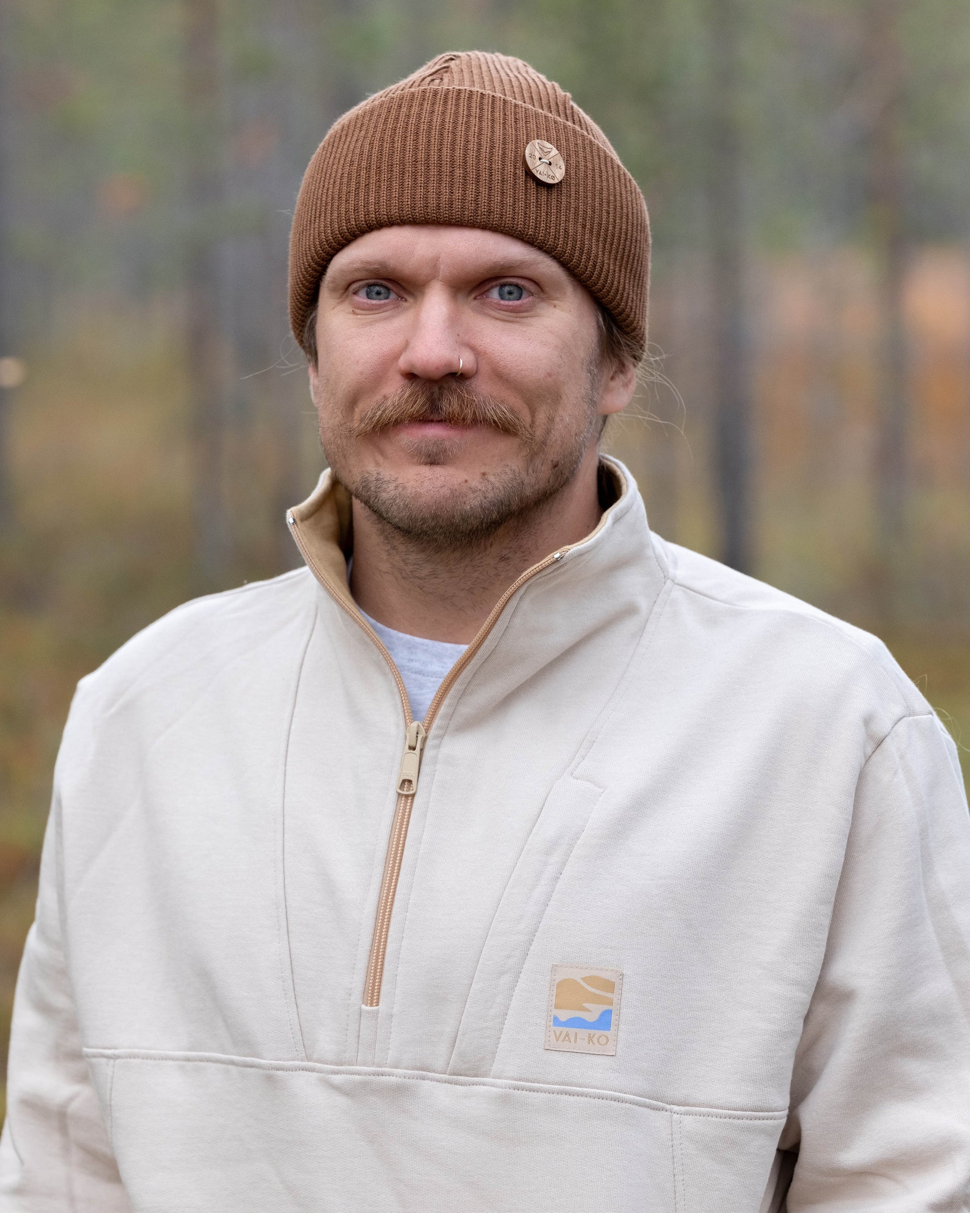 Man with a brown beanie and beige pullover standing outside in a forested area, looking at the camera.