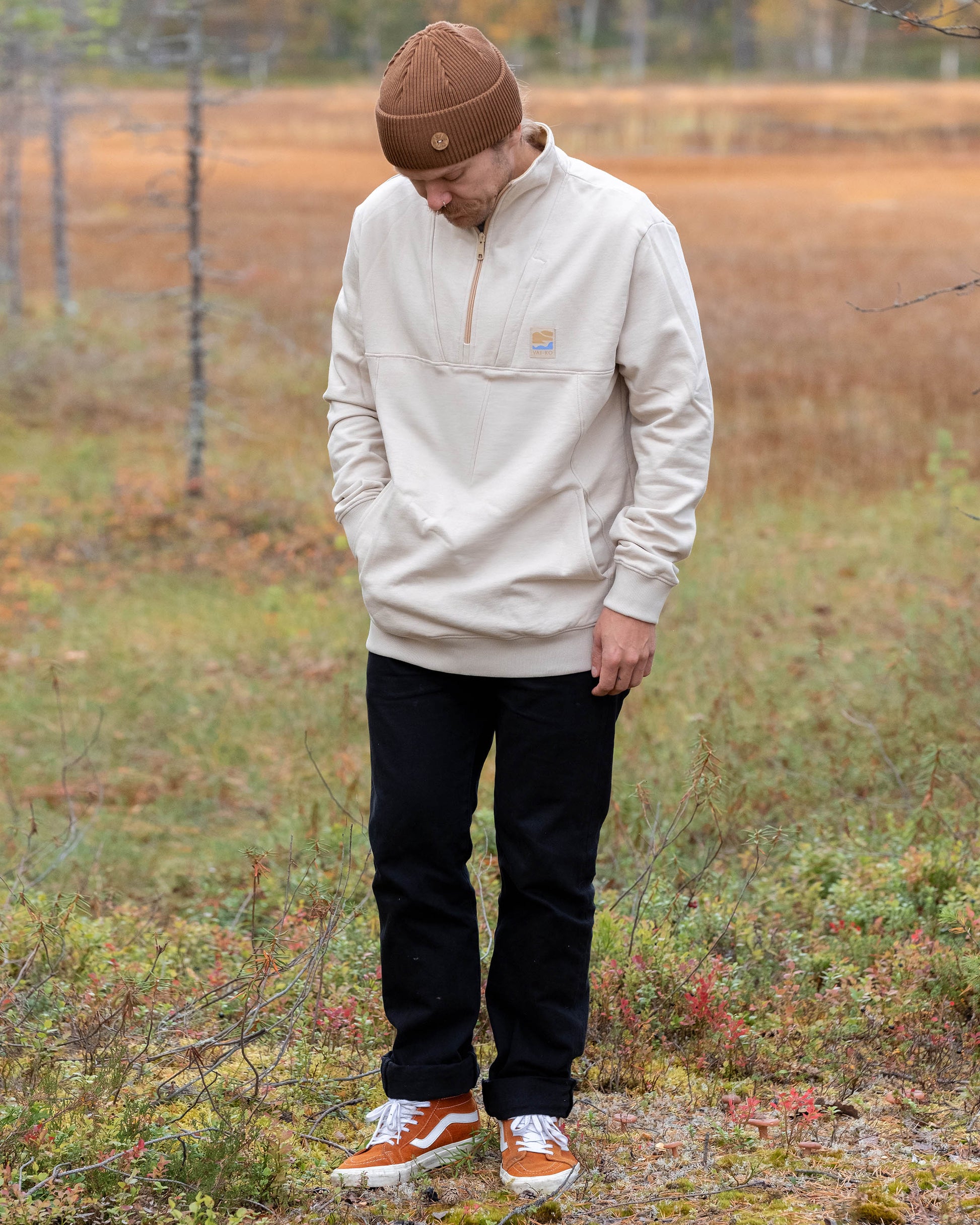 Man in beige sweater, black pants, and brown beanie standing outdoors looking down at the grass.