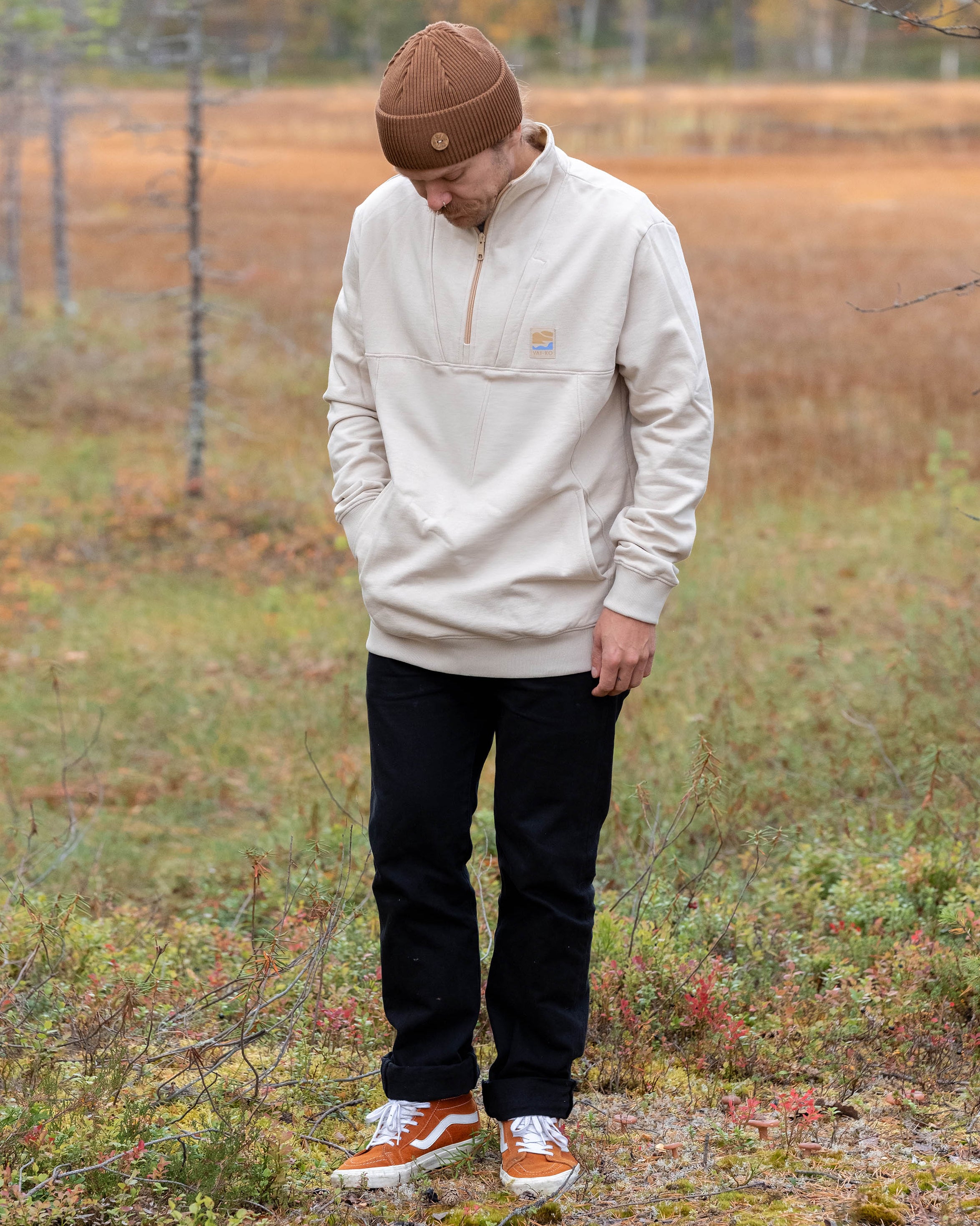 Man in beige sweater, black pants, and brown beanie standing outdoors looking down at the grass.